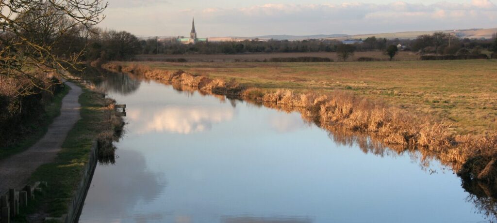 This view of the Chichester Canal features brown grass, small sidewalks, and a large open lot, highlighting peaceful outdoor activities and a few things to do in Hunston.